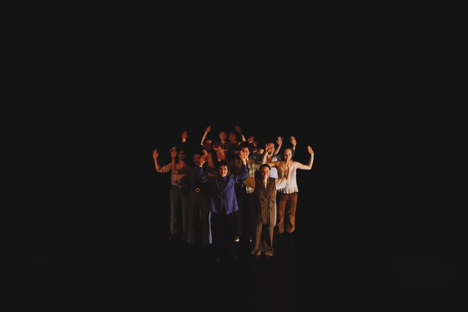 A distant cluster of performers with their arms raised on a black stage