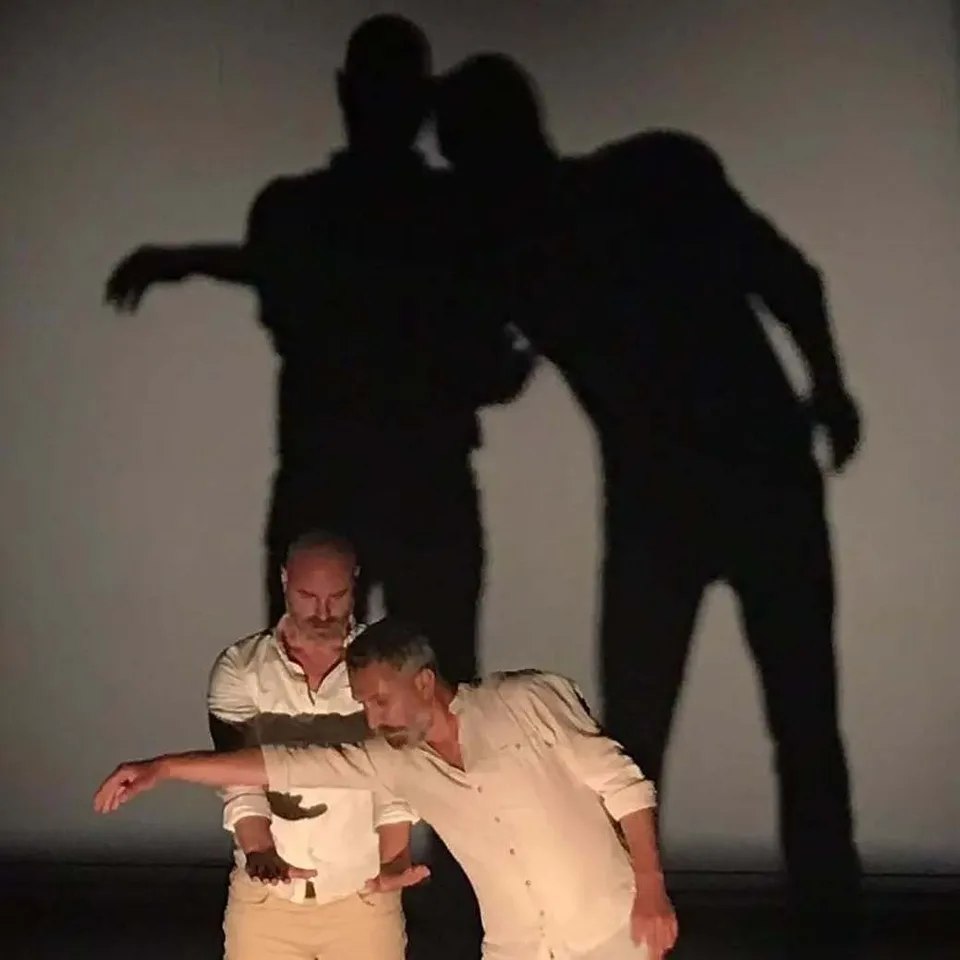 Two performers in pale clothes with their shadows thrown huge on the wall behind