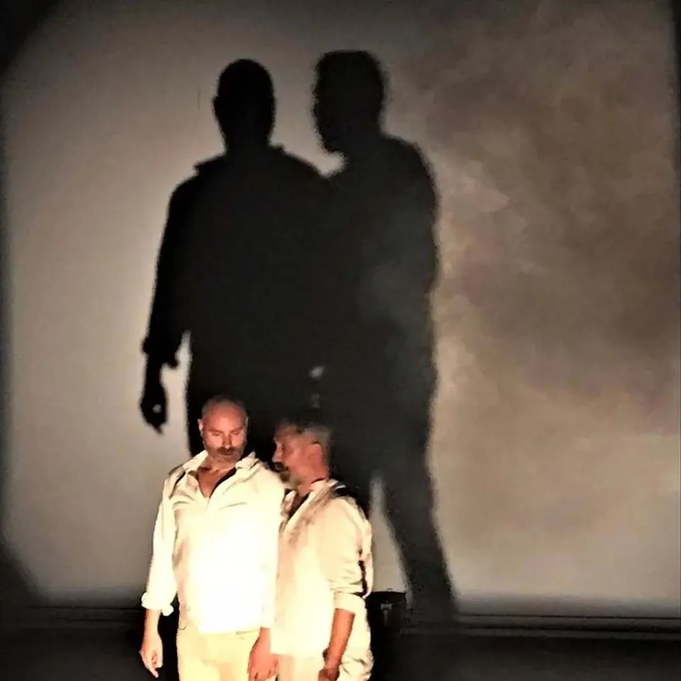 Two performers standing close together, two dark shadows looming over them
