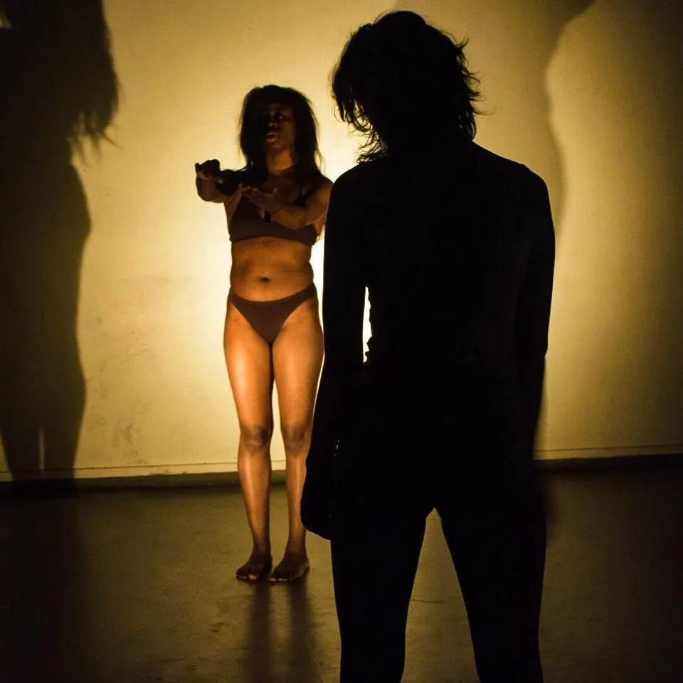 A performer in the light beside a tall dark silhouette, shadows spread across the wall