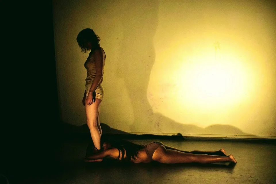 One performer stands over another lying down, against a glowing wall