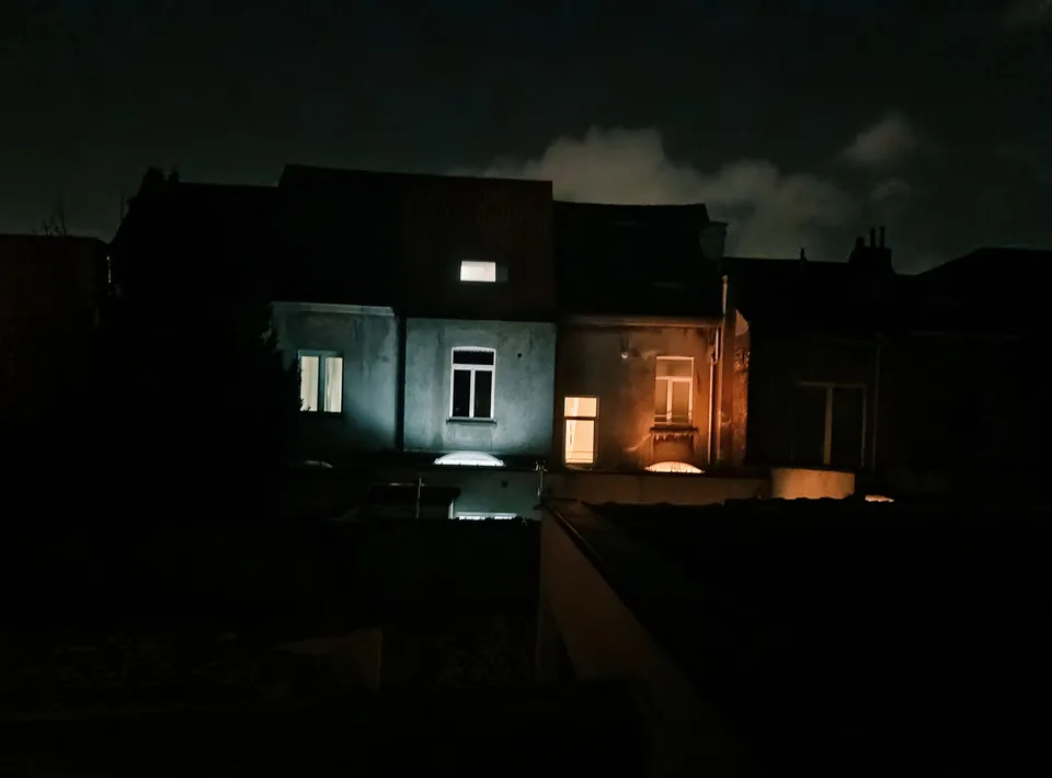 A dark suburban house at night, one window glowing yellow behind a hedge