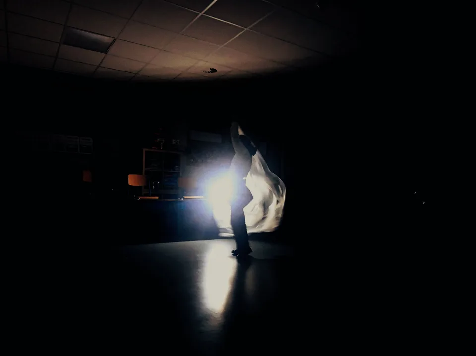 A figure caught in a shaft of white light in an otherwise black room, reflected in the floor