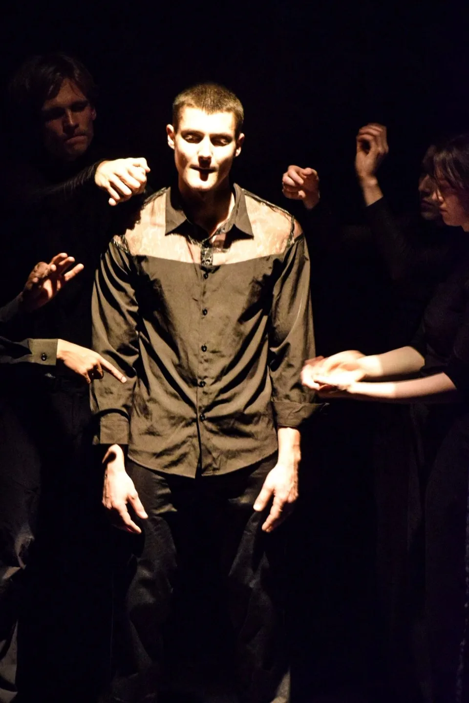 A performer lit at the centre while hands reach towards him out of the dark