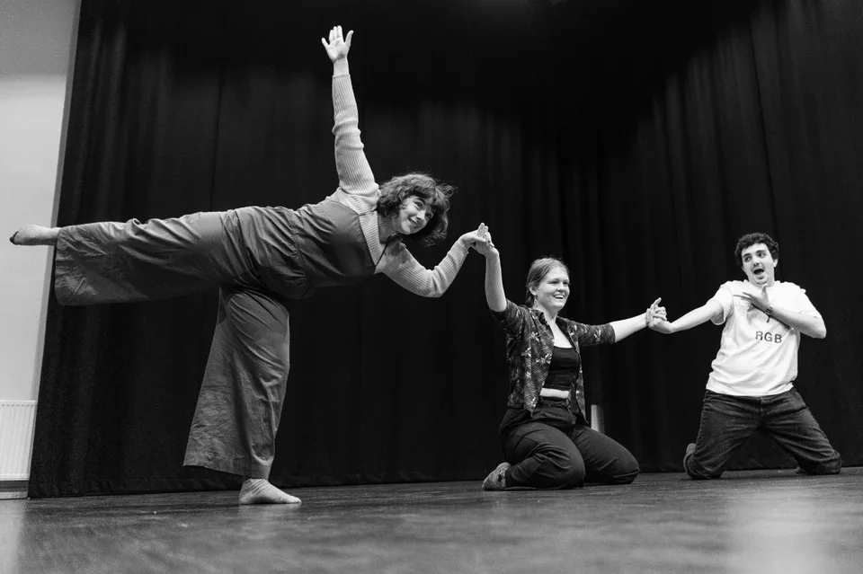 Black-and-white: a performer arched backwards on one leg while two others reach from the floor