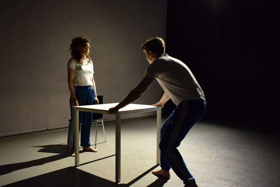 One performer stands at the table while the other pushes it across the floor
